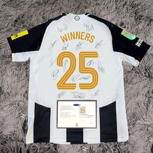 May include: White and black soccer jersey with the number 25 in gold and the word "WINNERS" above it. The jersey has black sleeves and stripes down the sides. Multiple signatures are visible on the jersey. A framed certificate is at the bottom.