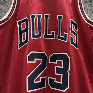Peut inclure: Un maillot de basket-ball rouge en tissu maill&eacute; arborant le mot BULLS en lettres noires avec des contours blancs arqu&eacute;s sur la poitrine. Sous le texte, le num&eacute;ro 23 est affich&eacute; en grands chiffres noirs avec des contours blancs sur le devant.