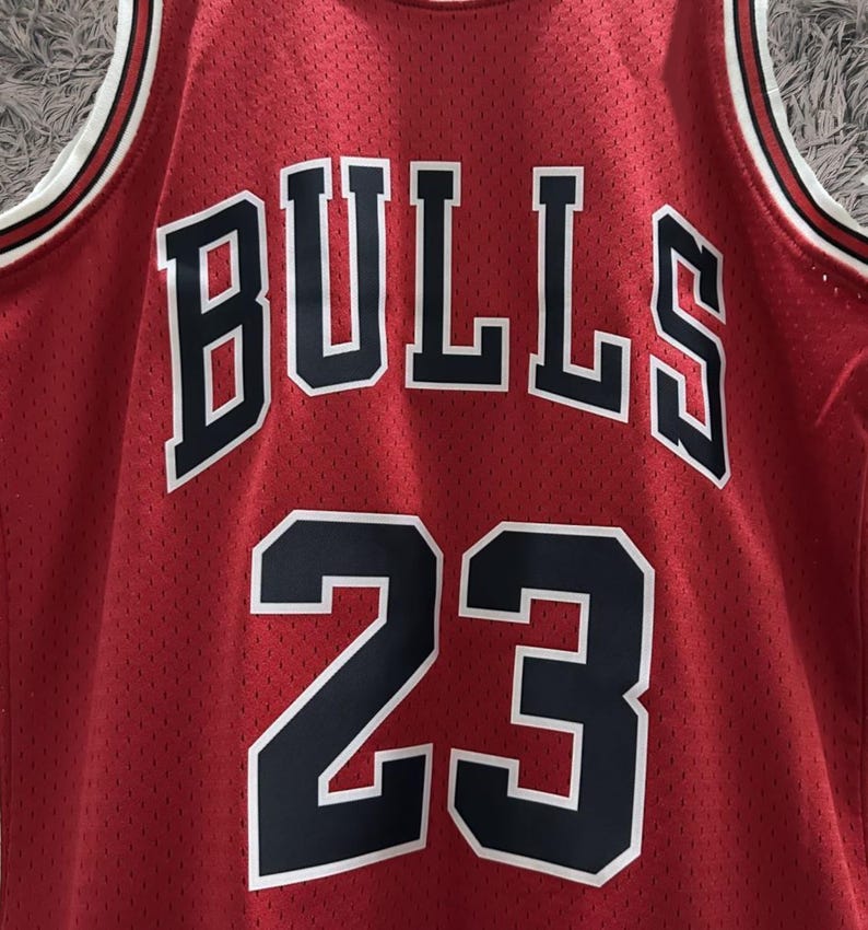 Peut inclure: Un maillot de basket-ball rouge en tissu maill&eacute; arborant le mot BULLS en lettres noires avec des contours blancs arqu&eacute;s sur la poitrine. Sous le texte, le num&eacute;ro 23 est affich&eacute; en grands chiffres noirs avec des contours blancs sur le devant.