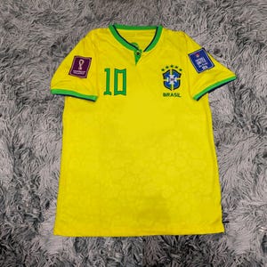 May include: A bright yellow short-sleeved jersey featuring a green collar, the number 10 in green on the chest, and the Brazil national team crest with five stars. The sleeves have green trim, and patches are visible on both shoulders.