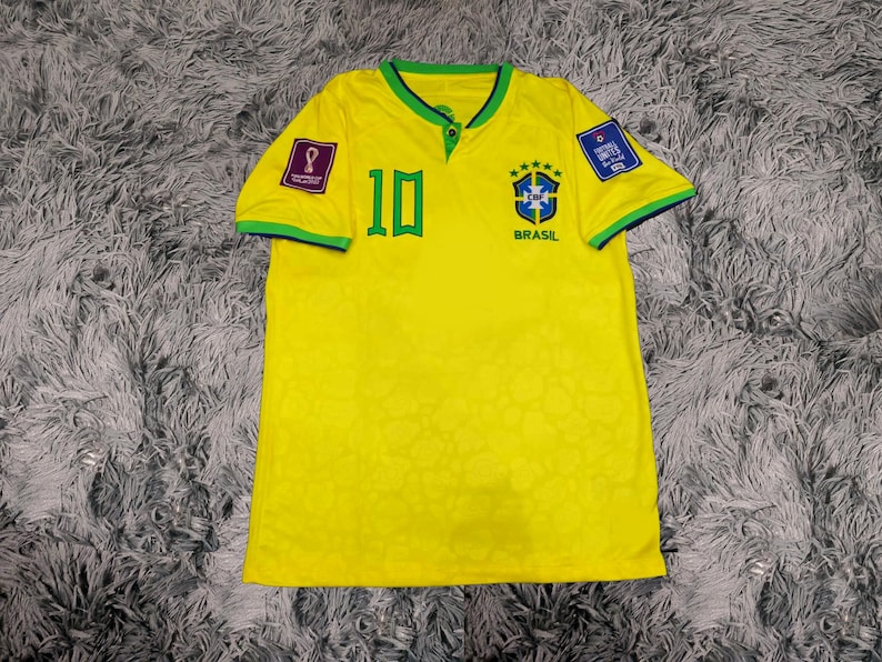 May include: A bright yellow short-sleeved jersey featuring a green collar, the number 10 in green on the chest, and the Brazil national team crest with five stars. The sleeves have green trim, and patches are visible on both shoulders.