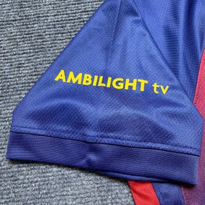 May include: Close-up of a blue athletic shirt sleeve with the text "AMBILIGHT tv" in yellow. The sleeve has a textured pattern and a dark blue trim. Red fabric is visible on the right side.