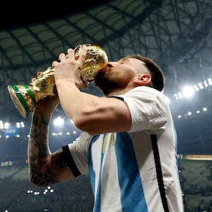May include: A person kissing a gold trophy with a green base. The trophy is held up with both hands. The person is wearing a white and light blue striped shirt. The background is a stadium.