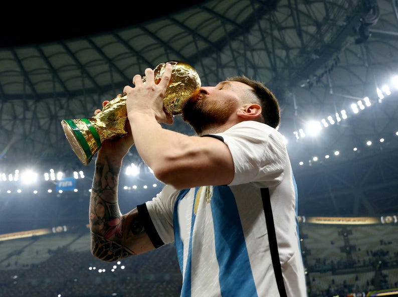 May include: A person kissing a gold trophy with a green base. The trophy is held up with both hands. The person is wearing a white and light blue striped shirt. The background is a stadium.