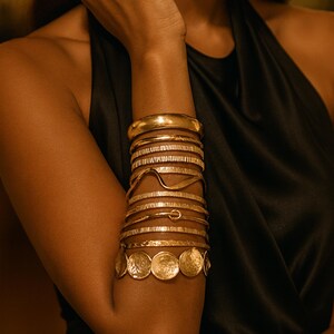 Könnte beinhalten: Eine Sammlung goldfarbener Armbänder in verschiedenen Designs schmückt einen Arm. Die Armbänder umfassen ein dickes, glattes Band, strukturierte Bänder und Bänder mit einzigartigen Formen und münzartigen Anhängern. Der Arm ist vor einem dunklen Hintergrund.