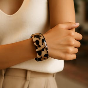 May include: A wide, tortoiseshell-patterned cuff bracelet. The bracelet features a glossy finish with a mix of tan and dark brown spots. The jewelry is worn on a wrist.