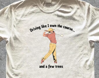 Pode incluir: T-shirt branco sujo com um design temático de golfe. A ilustração mostra um jogador de golfe em ação, com o texto: "Driving like I own the course... and a few trees."