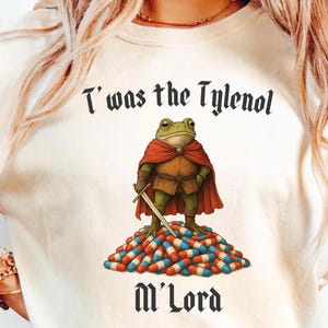 Pode incluir: Camiseta creme com um sapo de desenho animado com capa vermelha e espada, em pé sobre uma pilha de pílulas. O texto "T'was the Tylenol M'Lord" é impresso acima e abaixo da ilustração do sapo.