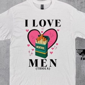 Può includere: T-shirt bianca con la scritta nera "I LOVE MEN (THOLS)". Un cuore rosa con contorno nero e un pacchetto di sigarette verde e giallo con la scritta "MENTHOL" sono al centro. Piccoli cuori e stelle rosa circondano il disegno.