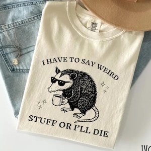 May include: Ivory-colored t-shirt with the text "I HAVE TO SAY WEIRD STUFF OR I'LL DIE" and a graphic of a possum wearing sunglasses and holding a mug. The shirt is folded on top of blue jeans, a tan hat, and a necklace.