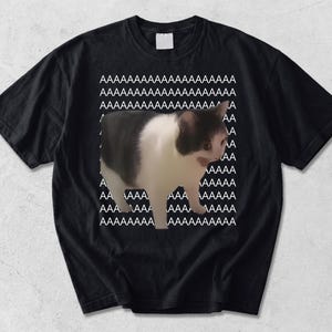 Könnte beinhalten: Ein schwarzes kurzärmeliges T-Shirt mit einer Grafik einer weiß-grauen Katze mit offenem Maul. Hinter der Katze ist der Buchstabe A in weißem Text in mehreren Zeilen wiederholt, was ein Hintergrundmuster erzeugt. Das T-Shirt hat einen Rundhalsausschnitt.
