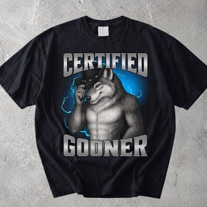 Puede incluir: Una camiseta negra de manga corta con un gráfico de un lobo musculoso con sombrero de vaquero. El texto "CERTIFIED" aparece sobre el lobo y "GOONER" debajo en letras plateadas metálicas, sobre un fondo de rayos de color azul brillante.