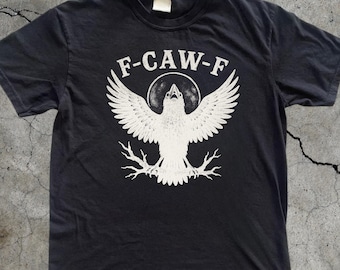 Pode incluir: Camiseta preta com um gráfico branco de um pássaro com as asas abertas, o texto "F-CAW-F" acima e a palavra "BLACK" na parte inferior. Um ícone de envio rápido está no canto superior direito.