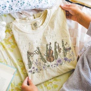 Op de afbeelding: Een ivoorkleurig T-shirt met korte mouwen en een grafische illustratie van vijf biddende sprinkhanen die muziekinstrumenten bespelen, waaronder een viool, fluit, cello, saxofoon en drums, omringd door kleine paarse bloemen en muzieknoten.