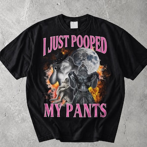 Może przedstawiać: Czarny t-shirt z krótkim rękawem z nadrukiem graficznym przedstawiającym wilka w kowbojskim kapeluszu oraz wilkołaka wyjącego do pełni księżyca na tle pomarańczowych płomieni. Napis I JUST POOPED MY PANTS jest wydrukowany różowymi literami.