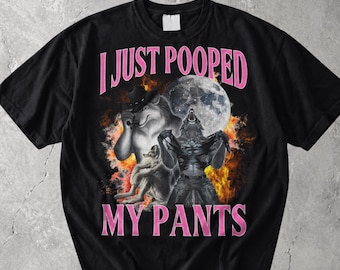 May include: A black short-sleeve t-shirt featuring a graphic print of a wolf wearing a cowboy hat and a werewolf howling at a full moon against a background of orange flames. The text I JUST POOPED MY PANTS is printed in pink capital letters.