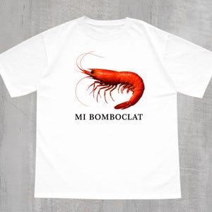 May include: A white short-sleeved t-shirt featuring a realistic orange-red shrimp graphic centred on the chest. Below the shrimp, the text MI BOMBOCLAT is printed in a simple black serif font. The shirt has a crew neckline and a standard fit.