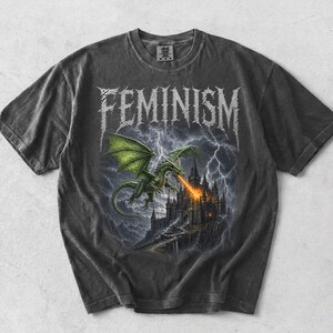 Può includere: Una t-shirt grigio antracite a maniche corte con la parola FEMINISM in un carattere in stile metallico nella parte superiore. La grafica raffigura un drago verde che vola sopra un castello scuro, sputando fuoco in un cielo tempestoso con fulmini.