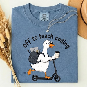 Può includere: Una t-shirt color blu jeans con la grafica di un'oca bianca con occhiali, su un monopattino, con uno zaino contenente un portatile e matite, e che tiene in mano un caffè. Il testo recita off to teach coding in caratteri neri.