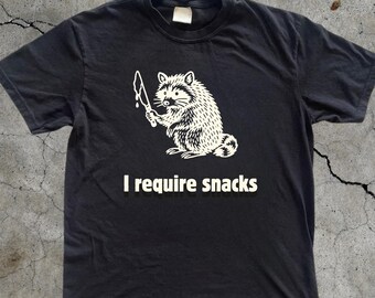 Pode incluir: Camiseta preta com um gráfico branco de um guaxinim segurando uma faca e o texto "I require snacks". A palavra "BLACK" está impressa na parte inferior. Uma peça de roupa casual.