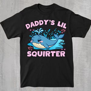 Könnte beinhalten: Ein schwarzes Kurzarm-T-Shirt mit einer Grafik eines blauen Wals, der Wasser spritzt. Der Text DADDY'S LIL SQUIRTER ist in Pink über und unter dem Wal gedruckt. Kleine pinke Herzen und blaue Wasserspritzer umgeben die zentrale Wal-Illustration.