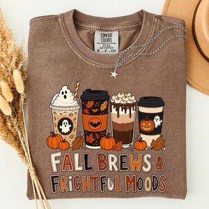 Op de afbeelding: Een bruin t-shirt met ronde hals en een afbeelding van vier koffiebekers met Halloween-thema's, waaronder spoken, pompoenen en vleermuizen. De tekst "FALL BREWS & FRIGHTFUL MOODS" staat in oranje en witte letters. Voorzien van een Comfort Colors label.