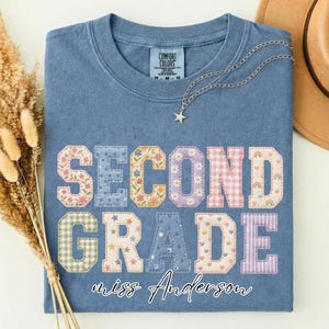 Può includere: Una t-shirt color blu jeans con scollo rotondo ed etichetta Comfort Colors. La parte anteriore presenta il testo SECOND GRADE in grandi lettere stampatello con motivi vari, con il nome miss Anderson scritto in corsivo bianco sotto.