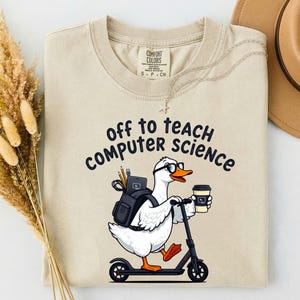 May include: An ivory t-shirt featuring a graphic of a white duck wearing glasses, carrying a black backpack with pencils and a tablet, riding a black scooter, and holding a coffee cup. The text reads "off to teach computer science".