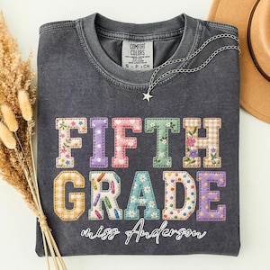Puede incluir: Una camiseta de cuello redondo gris carbón con el texto FIFTH GRADE en letras mayúsculas grandes y coloridas con patrones florales, de cuadros y de lunares. Debajo del texto, el nombre miss Anderson está escrito en cursiva blanca.