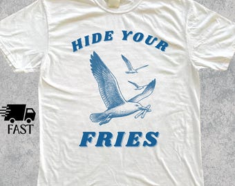 Pode incluir: Camiseta branca com um desenho azul de uma gaivota carregando batatas fritas e o texto "HIDE YOUR FRIES". Também há duas gaivotas menores em voo. O texto "COLOR: WHITE" está no topo.