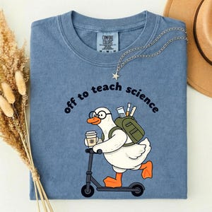 Può includere: Una t-shirt color blu jeans con la grafica di un'anatra bianca con occhiali, uno zaino verde con strumenti scientifici, che tiene una tazza di caffè e va su un monopattino. Il testo "off to teach science" è stampato sopra l'anatra.