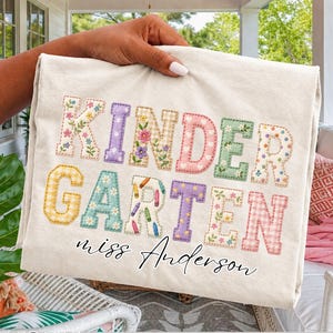 Può includere: Una t-shirt color avorio con la parola KINDERGARTEN in grandi lettere maiuscole multicolori, a fantasia e ricamate con fiori, con il nome miss Anderson stampato in corsivo nero sotto il testo principale.