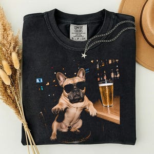 Könnte beinhalten: Ein schwarzes T-Shirt mit Rundhalsausschnitt und einem Grafikdruck eines beigen Französischen Bulldoggen mit schwarzer Sonnenbrille, der an einer Holztheke neben einem Glas Bier sitzt. Das Comfort Colors Etikett ist am Kragen sichtbar.