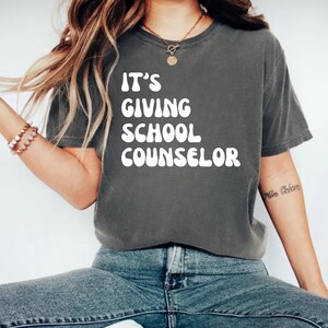 Può includere: Una maglietta grigio scuro con la scritta bianca "IT'S GIVING SCHOOL COUNSELOR" in stile retrò. La maglietta è abbinata a jeans blu. La parola "PEPPER" è in basso nell'immagine.