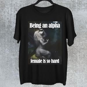 Puede incluir: Camiseta negra con la frase "Being an alpha female is so hard". El diseño muestra una criatura mítica con aspecto de lobo en un entorno forestal. La camiseta está colgada en una percha negra.
