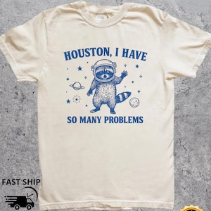 Può includere: T-shirt color crema con grafica blu di un procione con casco da astronauta, circondato da stelle e pianeti. Il testo recita: "Houston, I have so many problems."