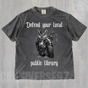 Può includere: T-shirt color pepe con la scritta "Defend your local public library". La grafica presenta un gufo in armatura con arco e frecce. La maglietta ha girocollo e maniche corte.