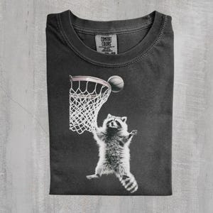 May include: Dark gray t-shirt featuring a raccoon playing basketball. The raccoon is reaching for the ball as it goes into the net. The shirt has a Comfort Colors tag.