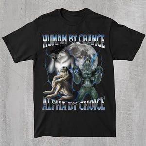 May include: Black t-shirt featuring a graphic design with three wolf-like creatures in front of a full moon. The text "HUMAN BY CHANCE" and "ALPHA BY CHOICE" are displayed in a blue and white font.