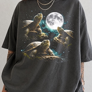 May include: A charcoal gray short-sleeve t-shirt featuring a graphic print of three turtles on rocks under a bright full moon in a starry night sky. The shirt has a crew neckline and a loose, relaxed fit with the word PEPPER printed in the corner.