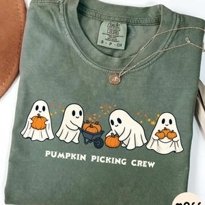 Può includere: T-shirt verde oliva con una grafica di quattro fantasmi dei cartoni animati che tengono zucche e la scritta "PUMPKIN PICKING CREW". La maglietta ha girocollo e maniche corte. Una collana d'argento con un ciondolo è drappeggiata sulla maglietta.