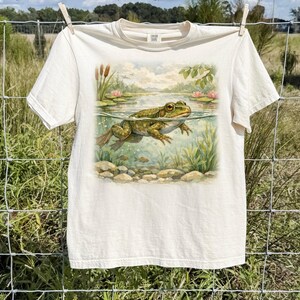 Può includere: T-shirt color avorio con un'illustrazione ad acquerello di una rana in uno stagno. Il design include ninfee, piante acquatiche e un paesaggio sereno. La maglietta è appesa a un filo.