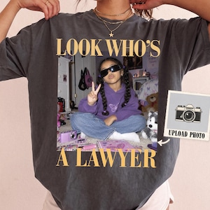May include: A charcoal grey t-shirt featuring a central photographic print of a child wearing sunglasses and a purple hoodie, making a peace sign. The text "LOOK WHO'S A LAWYER" is printed in large, light yellow capital letters above and below the image.