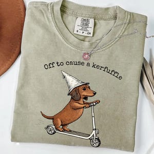 May include: Khaki-colored t-shirt featuring a brown dachshund wearing a wizard hat and riding a scooter. The text "Off to cause a kerfuffle" is printed above the dog. A silver necklace is draped across the shirt.