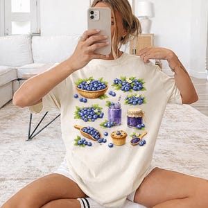 May include: Ivory t-shirt with a watercolour illustration of blueberries in a basket, sprigs, a smoothie, and a muffin. The word "IVORY" is printed at the bottom of the shirt.