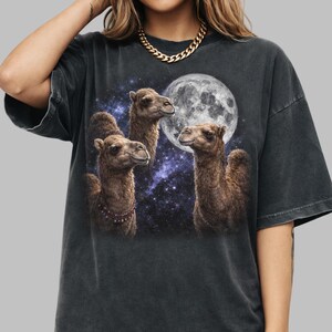 以下が含まれることがあります： 満月と星空を背景に、3頭のラクダが描かれたダークグレーのTシャツ。ラクダは茶色で毛並みが細かく、1頭はビーズのネックレスをしています。「PEPPER」の文字が下部にプリントされています。