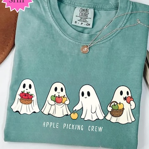 Könnte beinhalten: Hellgrünes T-Shirt mit Rundhalsausschnitt und kurzen Ärmeln. Das Shirt zeigt eine Grafik von vier Cartoon-Geistern, die Äpfel und Körbe halten, mit dem Text "APPLE PICKING CREW" darunter. Eine silberne Halskette mit Anhänger ist sichtbar.
