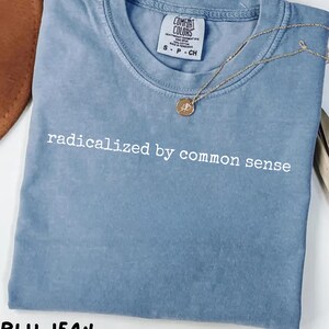 Op de afbeelding: Een blauw T-shirt met de witte tekst "radicalized by common sense". Het shirt heeft een ronde hals en korte mouwen. Een gouden ketting met een hanger is over het shirt gedrapeerd. Het shirt is gelabeld "Blu Jean".