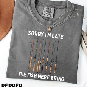 Può includere: T-shirt grigio con la scritta "SORRY I'M LATE THE FISH WERE BITING" e una grafica di canne da pesca. Una collana d'argento con un ciondolo è drappeggiata sulla maglietta. La maglietta è del marchio Comfort Colors.