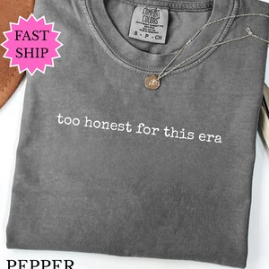 Puede incluir: Una camiseta gris con la frase "too honest for this era" en texto blanco. Un collar plateado con un pequeño colgante descansa sobre la camiseta. La camiseta está doblada y etiquetada como "Pepper".